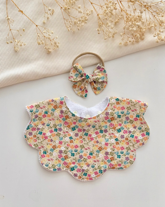 A baby bib and matching headband set with a colorful floral print on a cream background, styled on a light surface with dried flowers and a cream ribbed fabric backdrop.